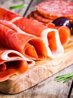 thin slices of prosciutto with olives and salami on wooden cutting board