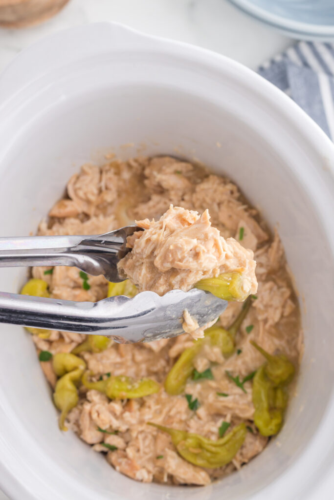 Shredded chicken in a Crock Pot. Tongs are lifting out some of the chicken. 